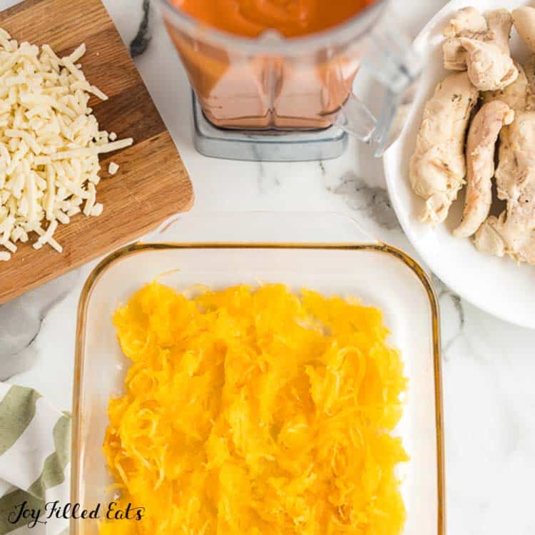 layer of spaghetti squash in casserole dish surrounded by a cutting board of shredded cheese, blender of pink sauce and white plate of chicken tenders