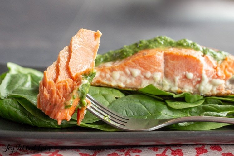 Fork with piece of salmon with pesto sauce on it. In front of salmon filet on a bed of spinach on black plate