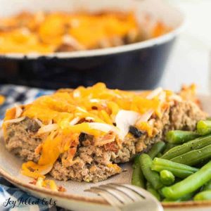 serving of burger bake and side of green beans on a plate with fork resting on edge of plate set in front of round casserole dish of burger bake