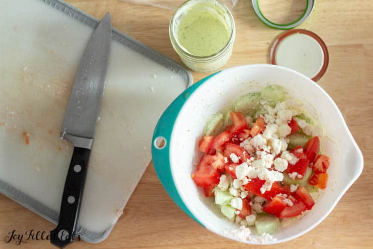 mixing bowl of cucumber and tomato salad ingredients next to a cutting board with knife and open mason jar of dressing