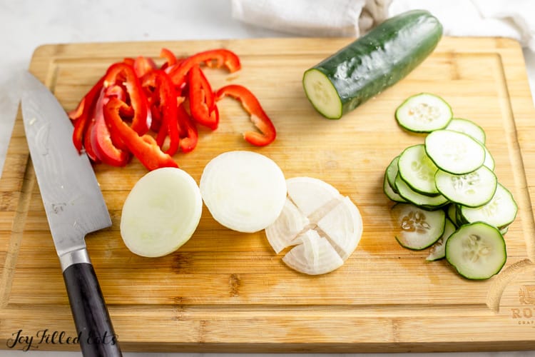 cutting board and knife of sliced keto antipasto salad ingredients including a red bell pepper, white onion and a cucumber