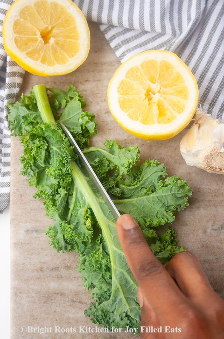 hand holding knife on kale leaf on cutting board to remove large, thick spine, next to lemon halves and garlic cloves