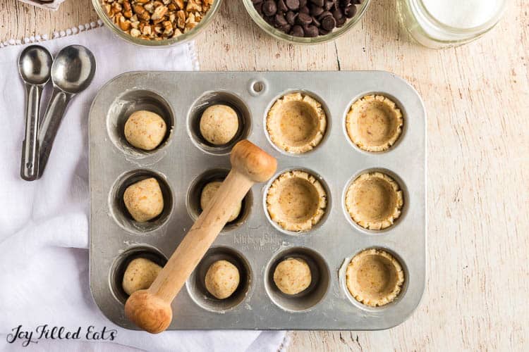 balls of tassie dough in muffin tin with wooden mallet shaping dough into small cups within muffin tin