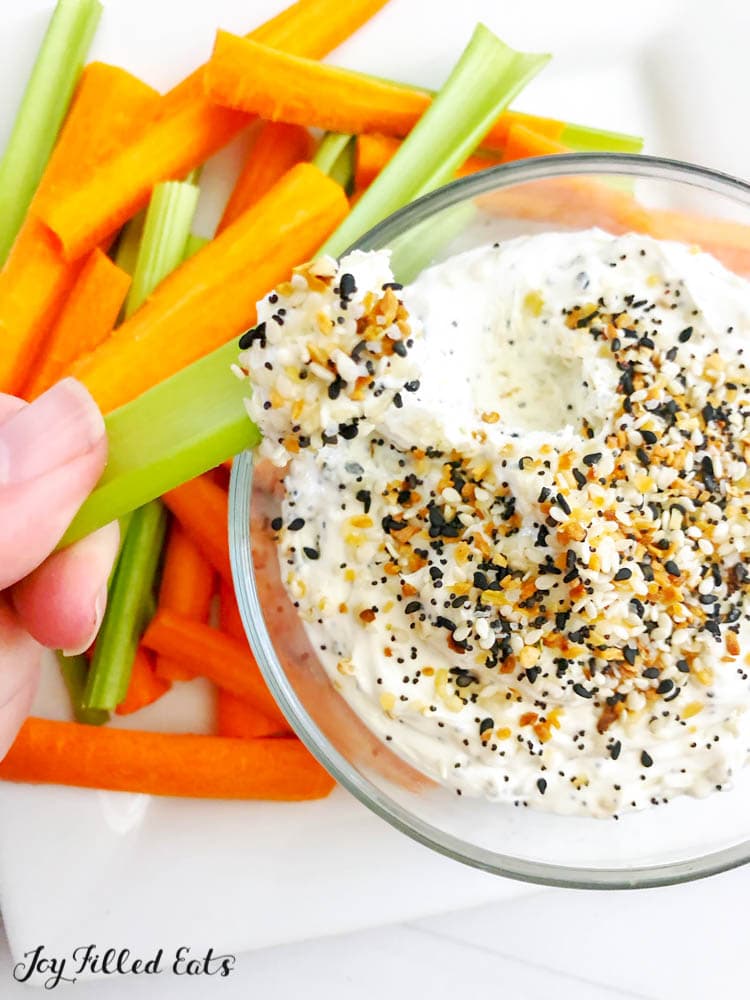 Hand holding celery stick with everything bagel dip on end in front of platter of celery and carrot sticks with a bowl of everything bagel dip