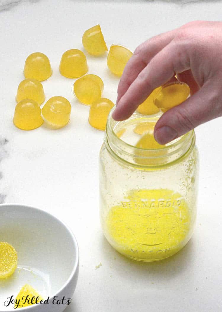 hand placing gumdrop into mason jar of sugar coating next to uncoated lemon gumdrops