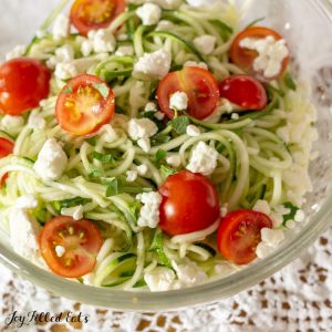 glass bowl of zucchini noodle salad topped with tomatoes and feta cheese on a white lace tablecloth