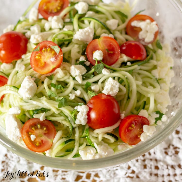 glass bowl of zucchini noodle salad topped with tomatoes and feta cheese on a white lace tablecloth