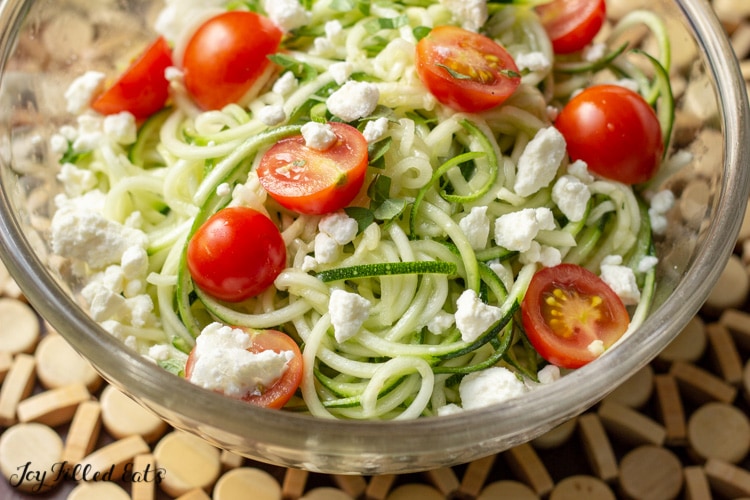 close up of zucchini salad in a glass bowl topped with feta cheese and tomatoes