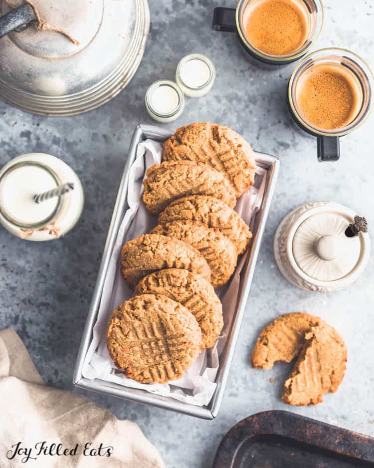 Flourless Peanut Butter cookies arranged in a parchment lined bread pan surrounded a halved cookie, two glass mugs of coffee, and other kitchen items