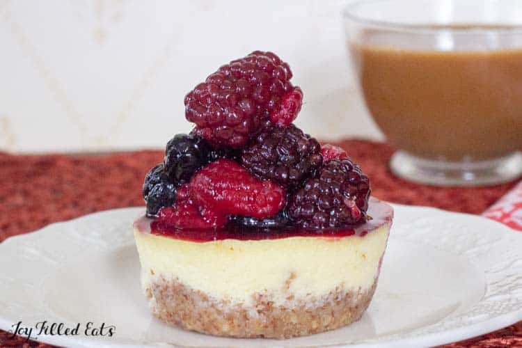 individual cheesecake topped with a pyramid of mixed berries on a white plate in from of a glass mug of coffee