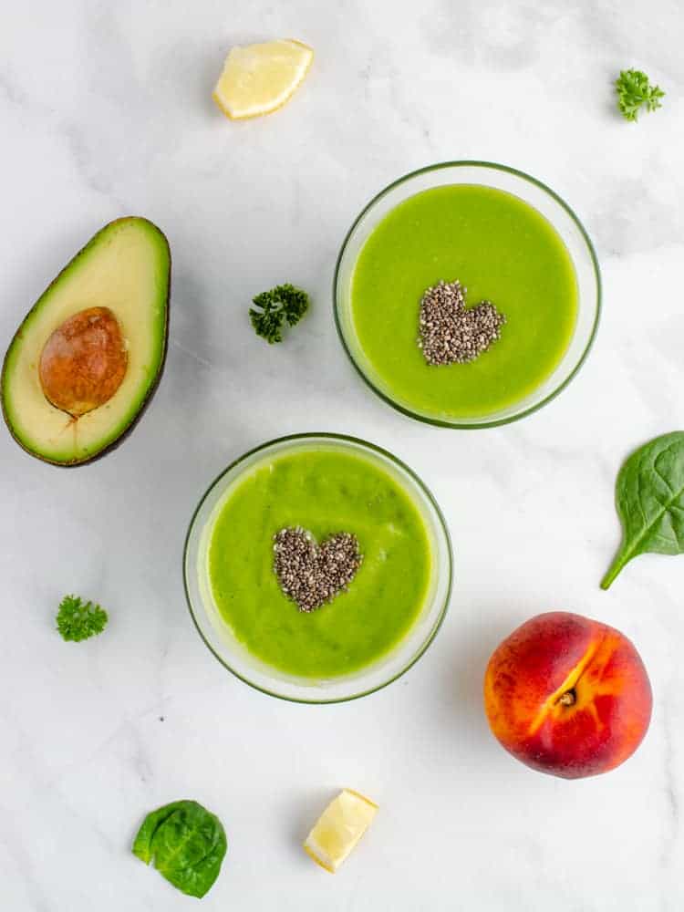 overhead view of two glasses of green smoothies topped with a seed shaped heart. Glasses are surrounded by various ingredients