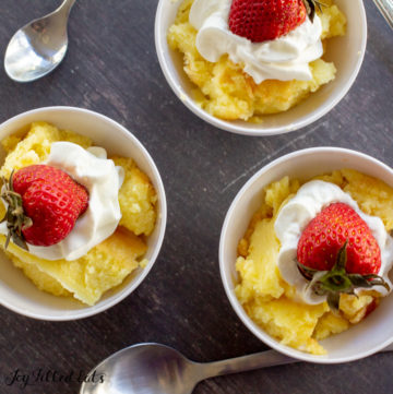 Overhead view of three small bowls of Baked Custard with Lemon each topped with whipped cream and a whole strawberry. Two spoons on the table below.