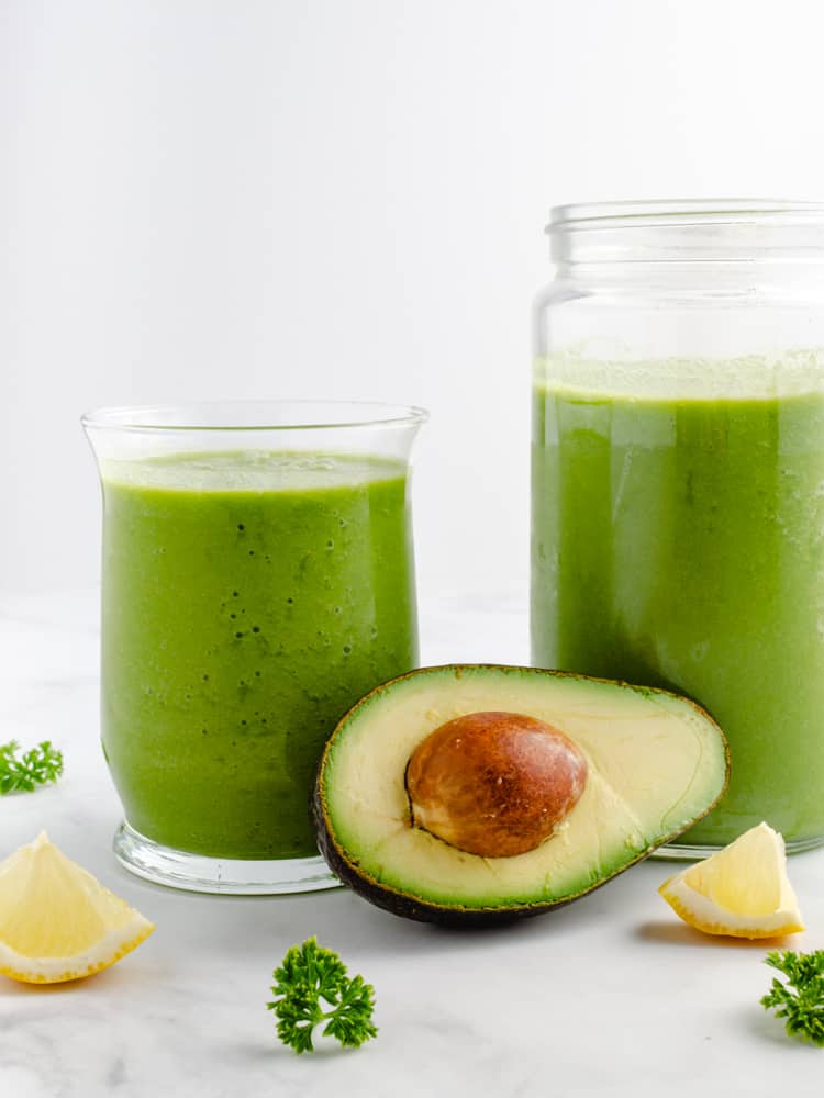 glass and jar of green smoothie with half avocado and pit in front