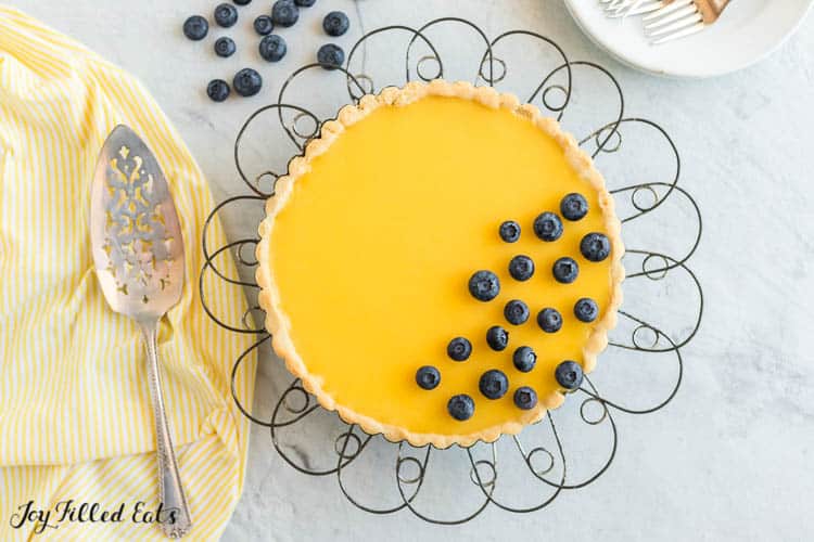 Lemon Curd Tart decorated with blueberries on a wire cake stand from above next to pie server on a decorative napkin
