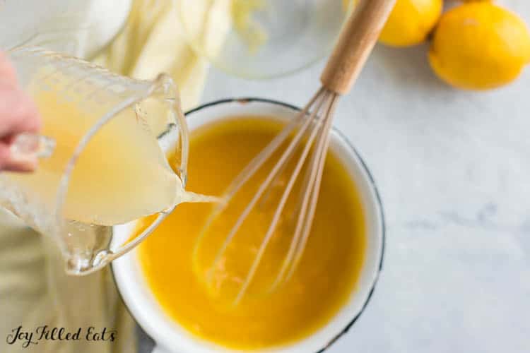 hand pouring measuring cup of lemon juice into saucepan with whisk for lemon curd