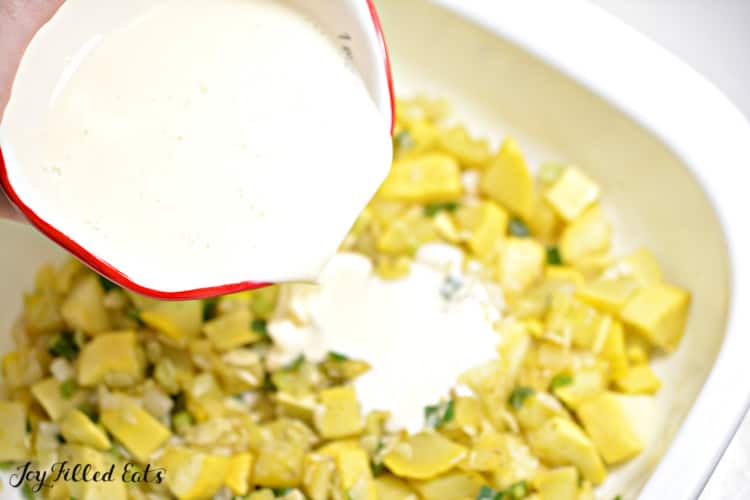 liquid ingredients for yellow squash casserole being poured into casserole dish filled with yellow squash pieces