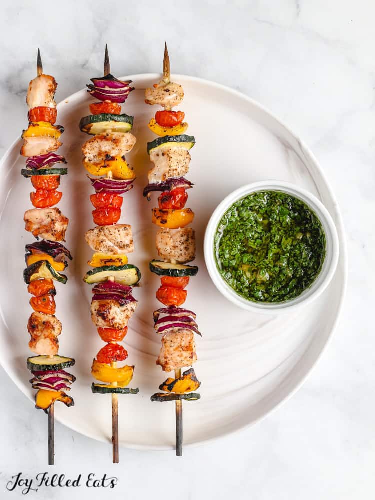 chicken skewers placed on a white plate next to a small dish of chimichurri sauce