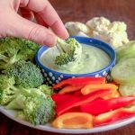 Hand dipping broccoli floret into small blue bowl of Creamy Chive Dip in center of veggie platter surrounded by Broccoli florets, Red Pepper slices, Cauliflower florets, and cucumber slices.