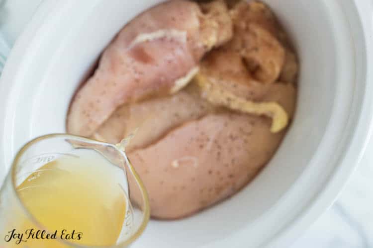 close up of measuring cup pouring liquid onto chicken breasts within crockpot
