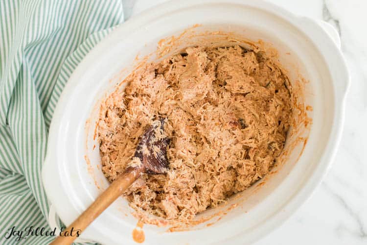 crockpot of shredded chicken being with seasoning with wooden spoon from above