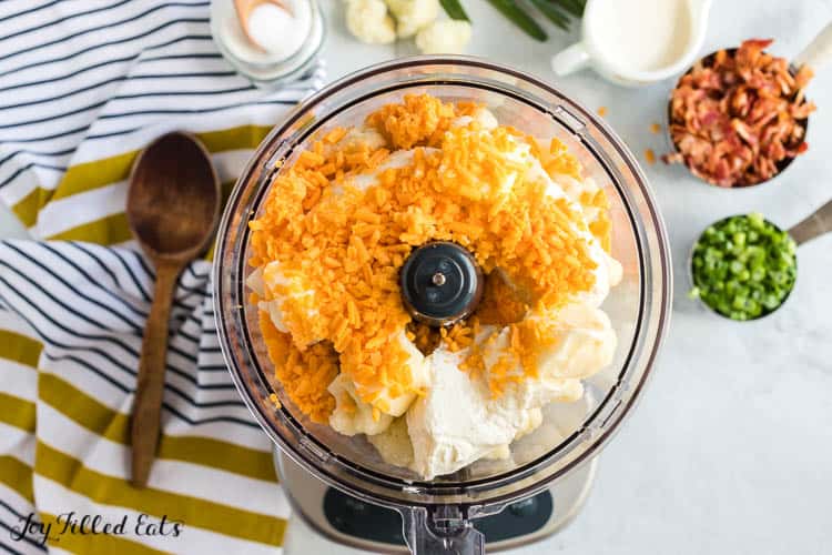 overhead view of food processor filled with loaded cauliflower casserole ingredients surrounded by a wooden spoon and measuring cups of other ingredients