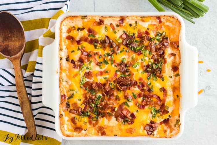 loaded cauliflower casserole topped with chives and bacon pieces in square white casserole dish set next to wooden spoon and striped napkin