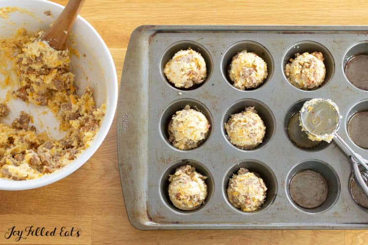 white mixing bowl of sausage muffin mixture with wooden spoon next to muffin tin and ice cream scoop. Muffin tin is half filled with small balls of sausage muffin mixture