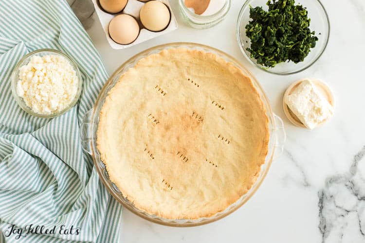 quiche crust in pie plate surrounded by bowl of other ingredients from above