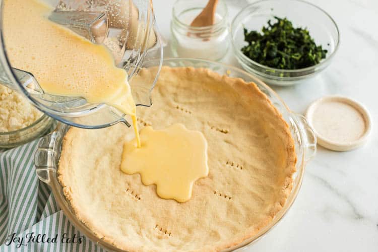 measuring cup pouring egg mixture into pie plate of quiche crust set next to a smaller dish of chopped spinach