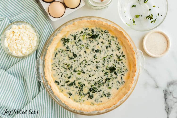 overhead view of pie plate filled with spinach and feta quiche filling poured over quiche crust. small dish of feta cheese crumbles is set nearby pie plate