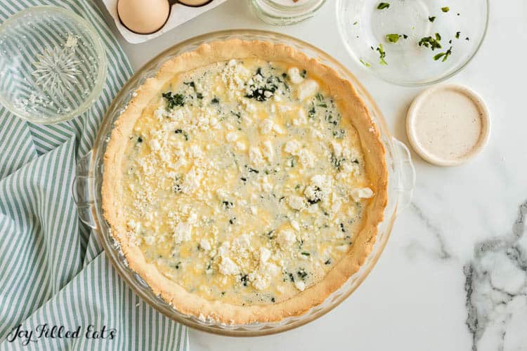 overhead view of pie plate filled with spinach and feta quiche filling topped with feta cheese crumbles all poured over quiche crust