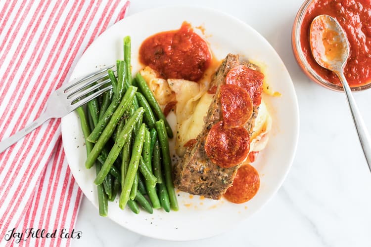 overhead view of fork and white plate with green beans, a slice of mozzarella stuffed meatloaf with cheese oozing onto plate topped with pepperoni and a small side of marinara sauce. next to plate is small dish of marinara sauce with a spoon
