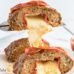 serving spatula lifting slice of pepperoni topped meatloaf from plate with melted cheese stretching from meatloaf slice to plate. Other meatloaf slices on plate have similar oozing melted cheese from center of loaf