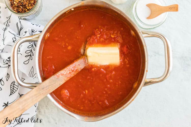 large pot of vodka sauce ingredients being mixed by a wooden spoon. portion of a stick of butter is set center, having just been added to the pot