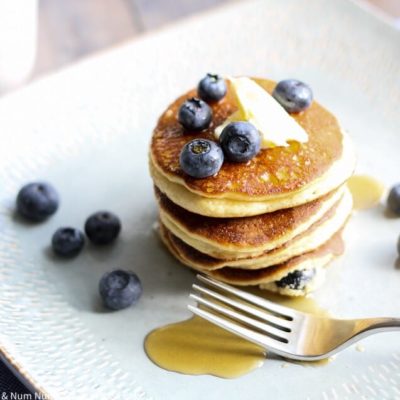 blueberry ricotta pancakes stacked center on a square plate topped with a pad of butter and blueberries scattered on top and around plate. syrup is drizzled on pancakes and plate with fork resting next to pancakes