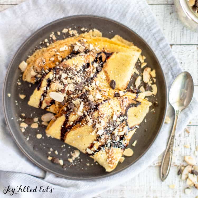 Keto crepes with drizzled chocolate sauce and topped with sliced almonds on a black plate, set on a cloth blue napkin table setting with spoon