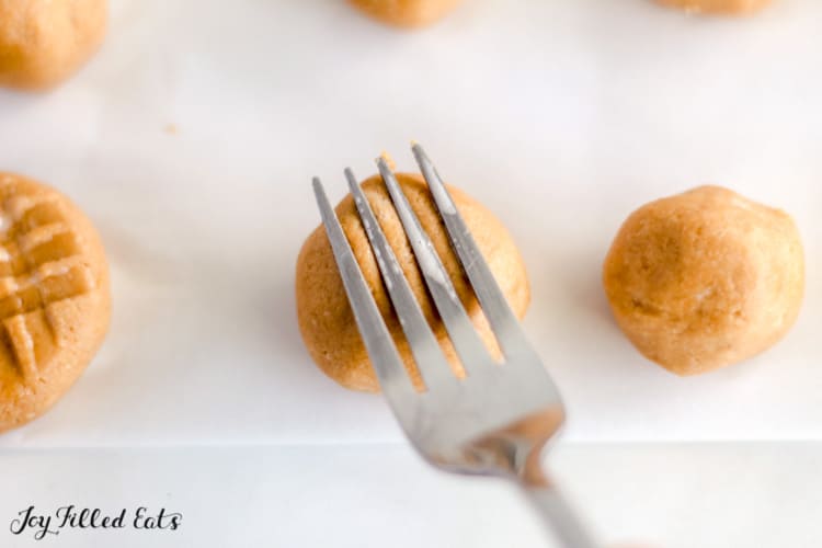 close up fork pressing indentations onto no bake peanut butter cookie ball surrounded by dough balls and shaped cookies on parchment paper