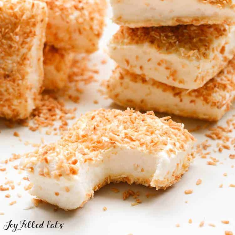 Close up of toasted coconut coated marshmallow with large bite taken out, in front of stacks of more toasted coconut coated marshmallows