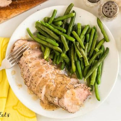 Pork Tenderloins slices fanned out on plate with fork served with a side of green beans. Tenderloin slices are dripped in gravy. Plate is set next to a decorative yellow napkin.