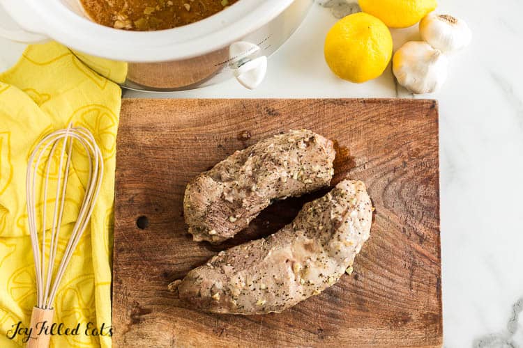 Two slow cooked pork tenderloin pieces on a cutting board placed next to a whisk, crock pot and lemons