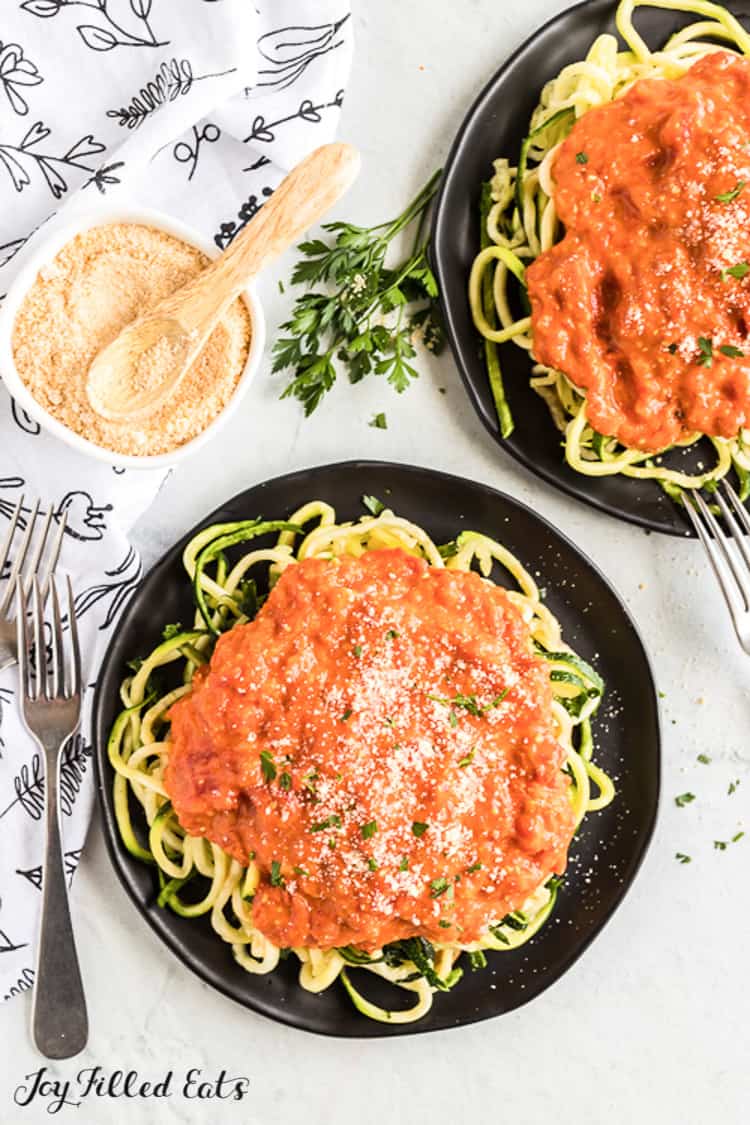 two black plates of zucchini noodles topped with a large amount of vodka sauce next to a small container of grated cheese and spoon