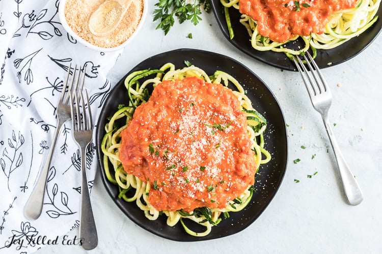 two black plates of zucchini noodles topped with a large amount of vodka sauce next to a couple forks and a small container of grated cheese and spoon. Vodka sauce is topped with grated cheese and parsley flakes