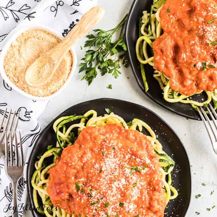 two black plates of zucchini noodles topped with a large amount of vodka sauce next to a small container of grated cheese and spoon