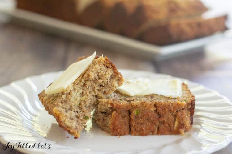 slice of low carb zucchini bread on a white plate torn in half. Each piece of bread is topped with a small spread of butter