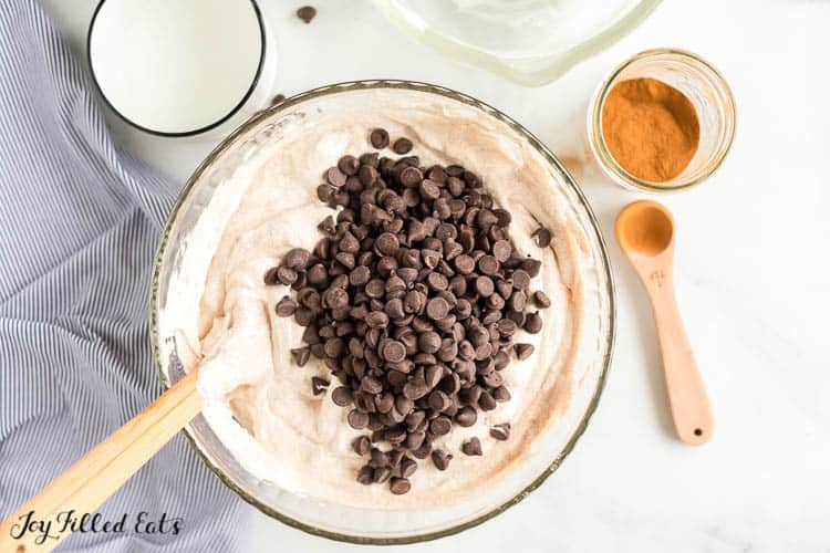large mixing bowl of cannoli cheesecake filling with chocolate chips poured on top ready to be mixed into filling with wooden handle spatula. Mixing bowl is next to small mason jar of cinnamon and small wooden teaspoon