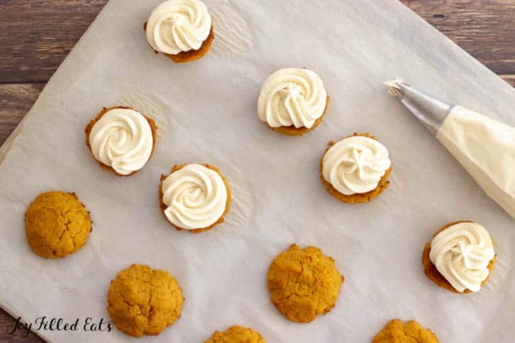 pumpkin sandwich cookies on parchment paper with icing piped on half the cookies along with full piping bag