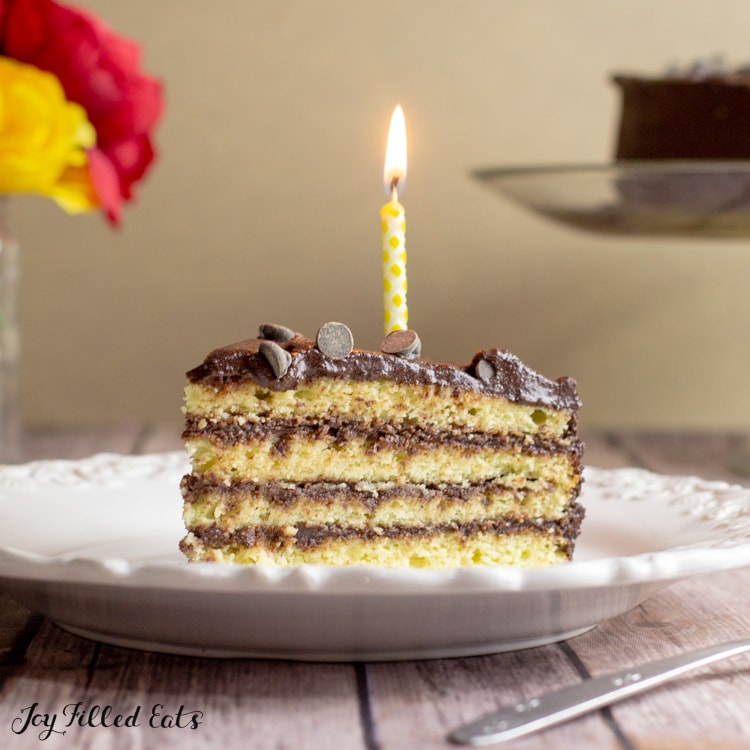 Slice of Keto Birthday cake layered with chocolate on a white plate. One lit yellow candle on the slice of cake.