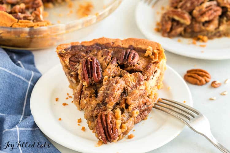 keto pecan pie slice on white plate with fork set in front of another slice of pie and pie plate with remaining pecan pie