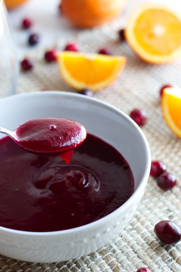 spoon full of cranberry jam mixture in front of large white bowl filled with jam. Surrounded by cranberries and orange wedges
