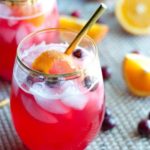 close up on glass and gold straw of vodka cranberry spritzer topped with ice, cranberries and orange peel